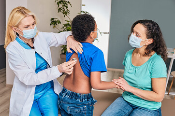 Doctor auscultating boy's back and lungs using a stethoscope. Child has pneumonia, mother wearing protective mask supports her son