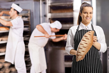 Woman wearing an apron and holding a loaf of brown bread and men working in a bakery