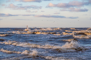 
Wavy water of the Baltic sea and cloudy sky in the evening. Orange Mangalsala lighthouse on the horizon. Gulls flying over the waves. Focus on foreground