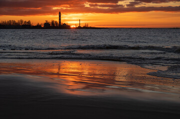 Naklejka na ściany i meble Beautiful feflections of orange sunset sky in seaside water in Mangalsala. Daugavgriva lighthouse with sun on the horizon in background. Focus on foreground