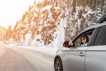 Happy smiling brunette sitting in a car and showing thumb up sign through opened side window in sunny winter day on icy road close up.
