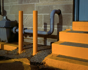 A minimalistic urban industrial geometric study of Bright yellow steps, protection poles, and a  gray gas meter at the rear door of the post office in Bllomington, Indiana 1986.