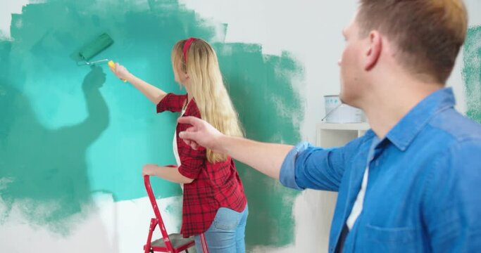 Portrait of beautiful young Caucasian woman stands on stepladder and painting wall in green color using roller brush while her husband looking and telling how to paint, renovation concept, home repair