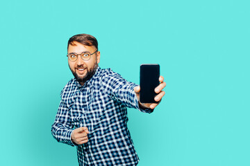 Image of joyful bearded man wearing glasses pointing finger on smartphone and expressing excitement on camera. isolated. blue background   