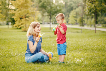 Naklejka na ściany i meble Young Caucasian mother and boy toddler son blowing soap bubbles in park. Mom and child playing having fun together outdoor on summer day. Happy authentic family childhood lifestyle.