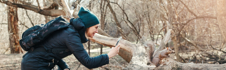 Woman taking picture photo of squirrel in park. Tourist traveler girl snapping smartphone photos of wild animal in forest. Fun outdoors activity and blogging vlogging online. Web banner header.