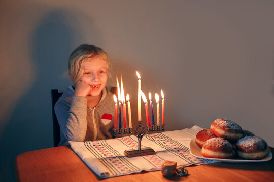 Girl lighting candles on menorah for traditional winter Jewish Hanukkah holiday at home. Child celebrating Chanukah festival of lights. Dreidel and Sufganiyot donuts on a table. Israel holiday.