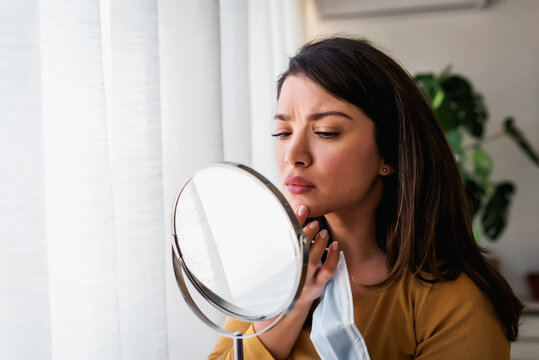 Woman Looking Herself In The Mirror At Home Standing By The Big Window. She Is Concerned About Acne, Maskne