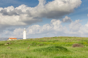 Fototapeta na wymiar White lighthouse on the background of the sky with clouds. Spring and lush grass, the sun is shining. Paphos, Cyprus.