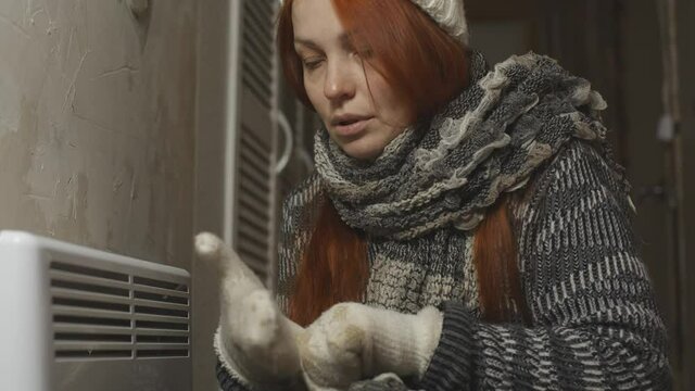 a woman is warming herself at the radiator in a cold house, problems with heating, heating the room with an electric convector