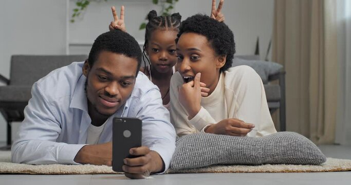 Loving happy afro american family taking photo on mobile phone camera smiling playing making faces for photography, father holding smartphone in his hands filming video while mom and daughter posing