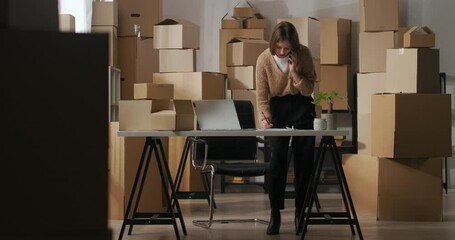 woman owner of small delivery company is communicating by phone with supplier in room of flat using as warehouse with boxes