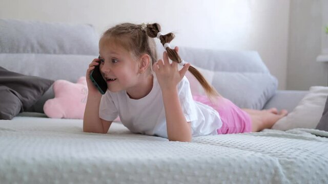 A 7-8 year old girl is lying on a gray sofa and talking on the phone.
The child communicates via a smartphone with friends.