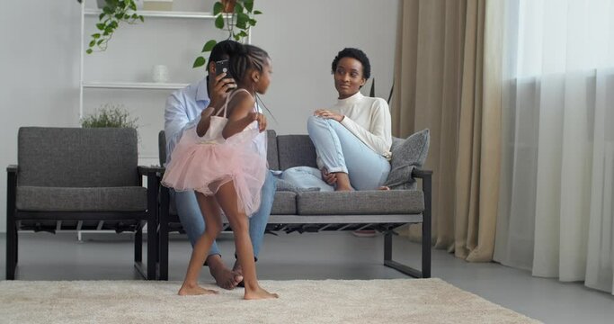 Young Afro American parents watch their beloved daughter dance, loving father makes video photo on smartphone camera of mobile phone while little girl princess ballerina dancing whirling in new dress