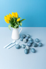 Jug with yellow tulips, Easter eggs, spoon and fork on blue background. Place for your text.