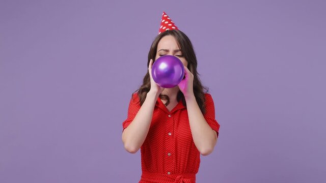 Funny cheerful young woman 20s in red dress birthday hat posing isolated on violet purple background in studio. Celebrating holiday party concept. Point index finger on air balloon inflating deflating