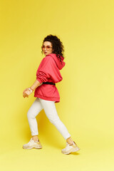 Fototapeta na wymiar A young woman in sportswear and sunglasses over a yellow background. Young beautiful woman in a pink hoodie and white pants at the yellow background, isolated