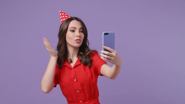 Funny young woman 20s in red dress birthday hat isolated on violet background studio. Celebrating holiday party concept. Doing selfie shot on mobile phone showing victory sign blowing sending air kiss