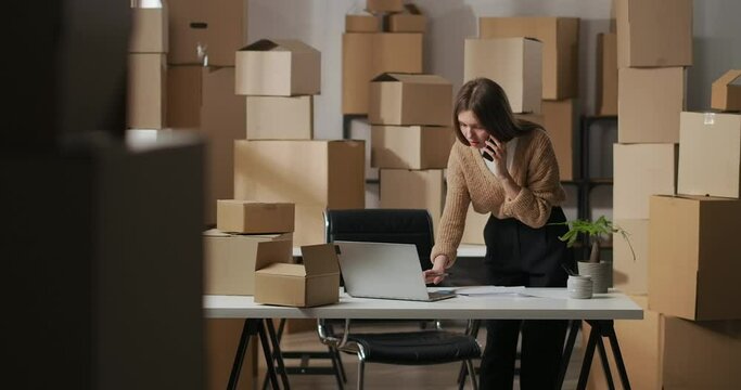 customer service of small delivery company, woman is consulting clients by phone and filling in order in laptop, portrait in home warehouse