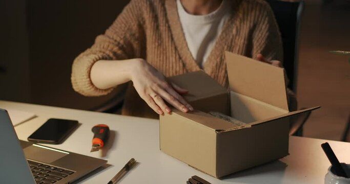 adult woman is packaging box with cosmetic for delivery, sitting at table at evening in room of apartment or office