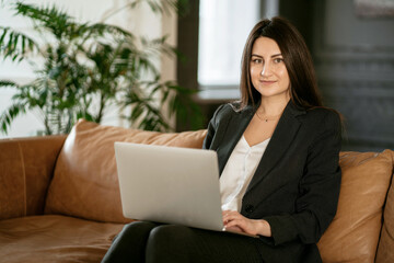 Naklejka premium The economist is a brunette woman. A lawyer employee works on a laptop computer. Business stylish black suit. Makes an order in an online store on a sofa in a modern office.