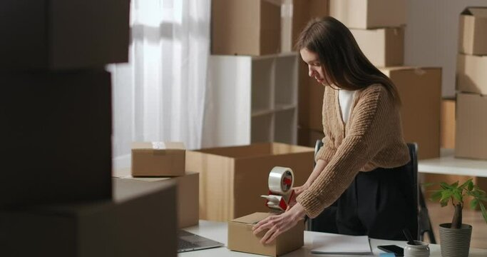 female worker of small domestic warehouse is using roll-on tape dispenser for packing box with goods, packaging products