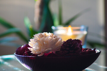 Bright white flower decoration in red bowl wth blurred bokeh scented candle in the background.