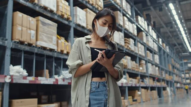 Young Asia businesswoman manager wear face mask looking for goods using digital tablet checking inventory levels stand in retail shopping center. Distribution, Logistics, Packages ready for shipment.