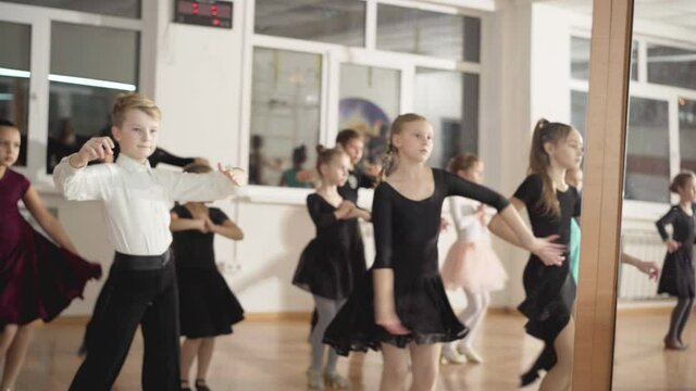 Reflection in mirror of group of children rehearsing latin ballroom movements with teacher walking. Talented Caucasian boys and girls practicing art performance in dancing school.