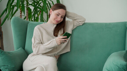 Smiling woman relaxes sitting on the couch at home using mobile phone text messages to chat with a friend