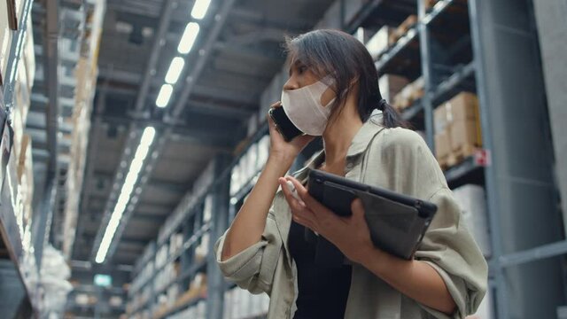 Young Asia businesswoman online seller wear face mask talking confirming order from customer on phone hold tablet stand in retail shopping center. Distribution, Logistics, Packages ready for shipment.
