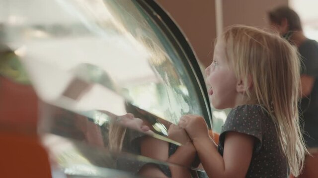 Medium shot of little Caucasian girl with blond hair wearing t-shirt standing at showcase in cafe and choosing ice-cream