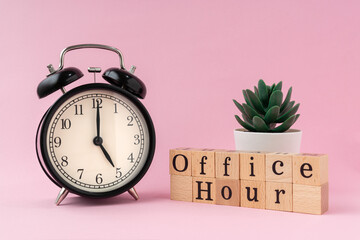 Black vintage alarm clock on a light pink background with a text office hour on wooden block. 