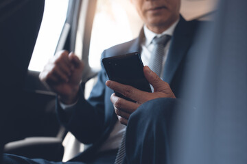 Businessman in black suit sitting on back seat inside a car and using mobile smartphone with morning sunlight
