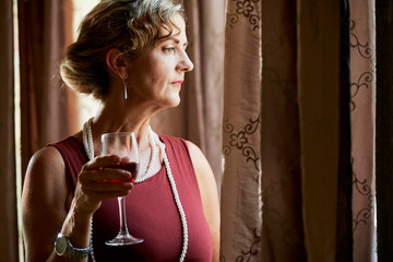 Elegant mature woman in maroon dress standing by window and drinking glass of wine