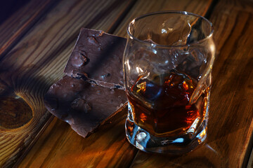 A glass of cognac and a bar of chocolate on a wooden table on a dark background