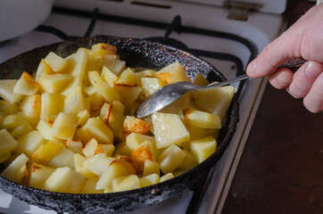 Man's Hand with a spoon stirs sliced potatoes in a pan on the kitchen stove.