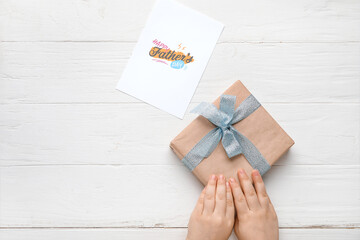 Hands with gift for Father's Day and greeting card on white wooden background