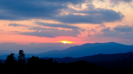 View of trees on a hill at cloudy sunset with fog light in the misty mountains at the background.