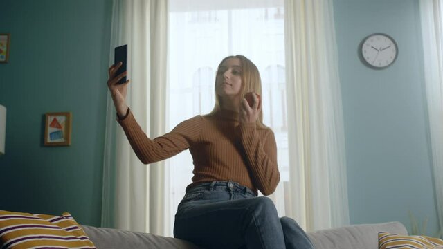 Young woman in blue jeans and brown sweater sits on back of sofa on background of window, holding red apple and taking selfie. Average plan.