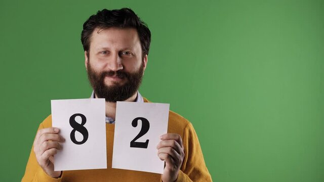 Man holding number 82 sign. Cheerful white bearded good-looking man in yellow sweater shows number eighty-two on white sheet. Chrome key, green screen, copy space