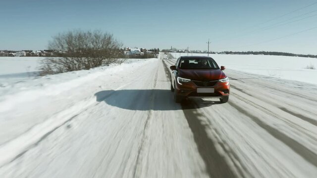 Low altitude aerial shot of a red car or SUV driving along rural snowy road in winter