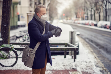 Businessman with shoulder bag wearing a face mask during the Covid-19 pandemic standing on a snowy urban street in winter checking the time on his wristwatch