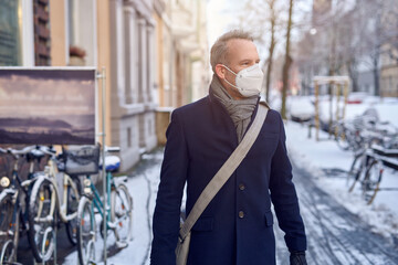 Man wearing a protective surgical face mask during the Covid-19 or coronavirus pandemic and winter overcoat with leather bag over his shoulder walking down a snowy urban street 