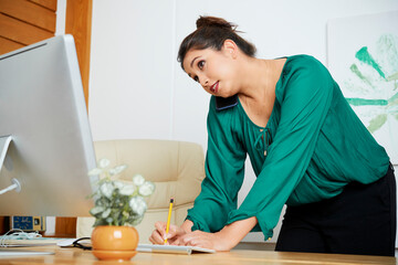 Pretty female entrepreneur leaning on office table, talking on phone with coworker or customer and writing in planner
