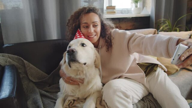 Young happy woman embracing cute golden retriever dog in party hat and taking selfie with smartphone while sitting on sofa at home