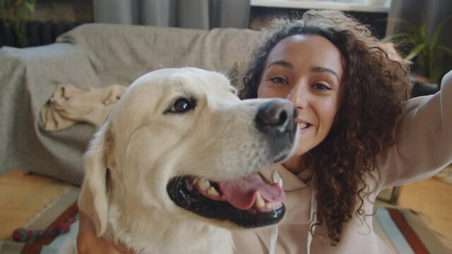 Young joyous woman holding smartphone in outstretched arm and posing for camera with adorable golden retriever dog while taking selfie at home