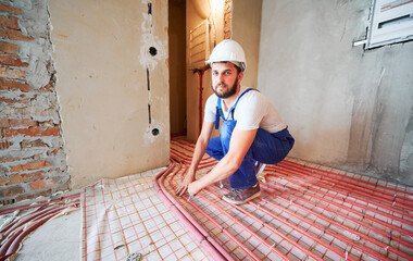 Male worker in safety helmet squatting and looking at camera while installing pipes for warm floor...