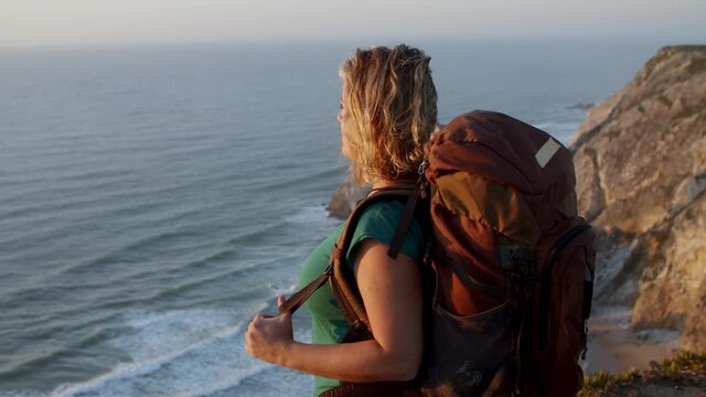 Happy middle aged woman wearing camping backpack, standing at rocky cliff, looking at breathtaking sea and sunset. Travel activity or freedom concept