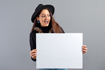 elegant young woman in glasses and hat showing empty board banner, isolated on gray, copy space